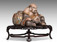 A finely painted japanese polychrome porcelain figure of hotei leaning against the bag of good fortune, l 22 cm - figure h 14,5 cm - total h 22,5 cm